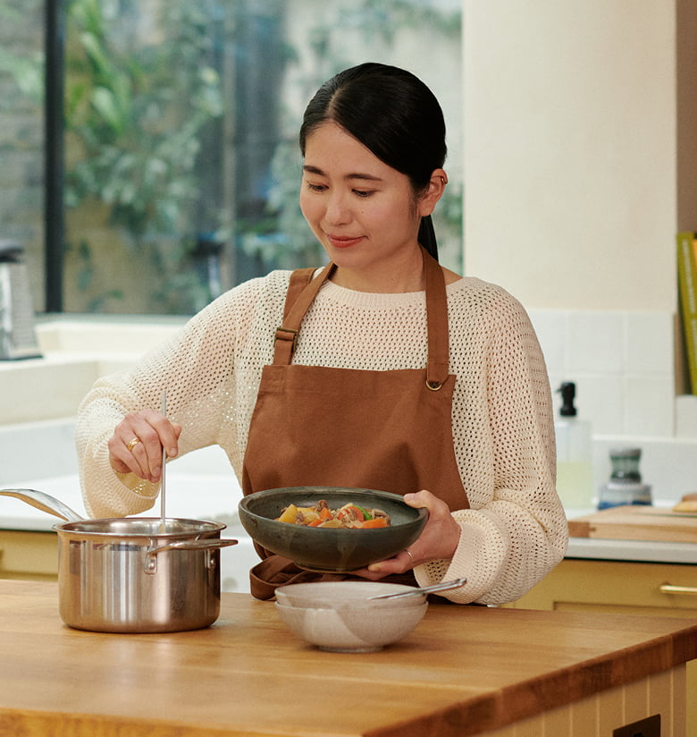 Photograph of a woman in a brown apron stirring a pot and holding a bowl of stew in a bright kitchen