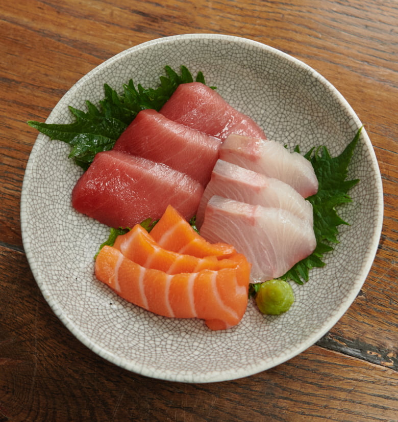Photograph of a bowl of assorted sashimi with tuna, salmon, and white fish on shiso leaves with wasabi